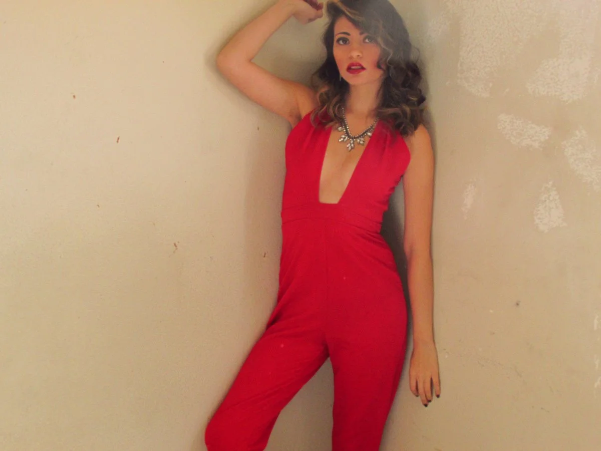 A woman in a red jumpsuit with a deep V neckline, holding her hair with one hand, standing against a beige wall.