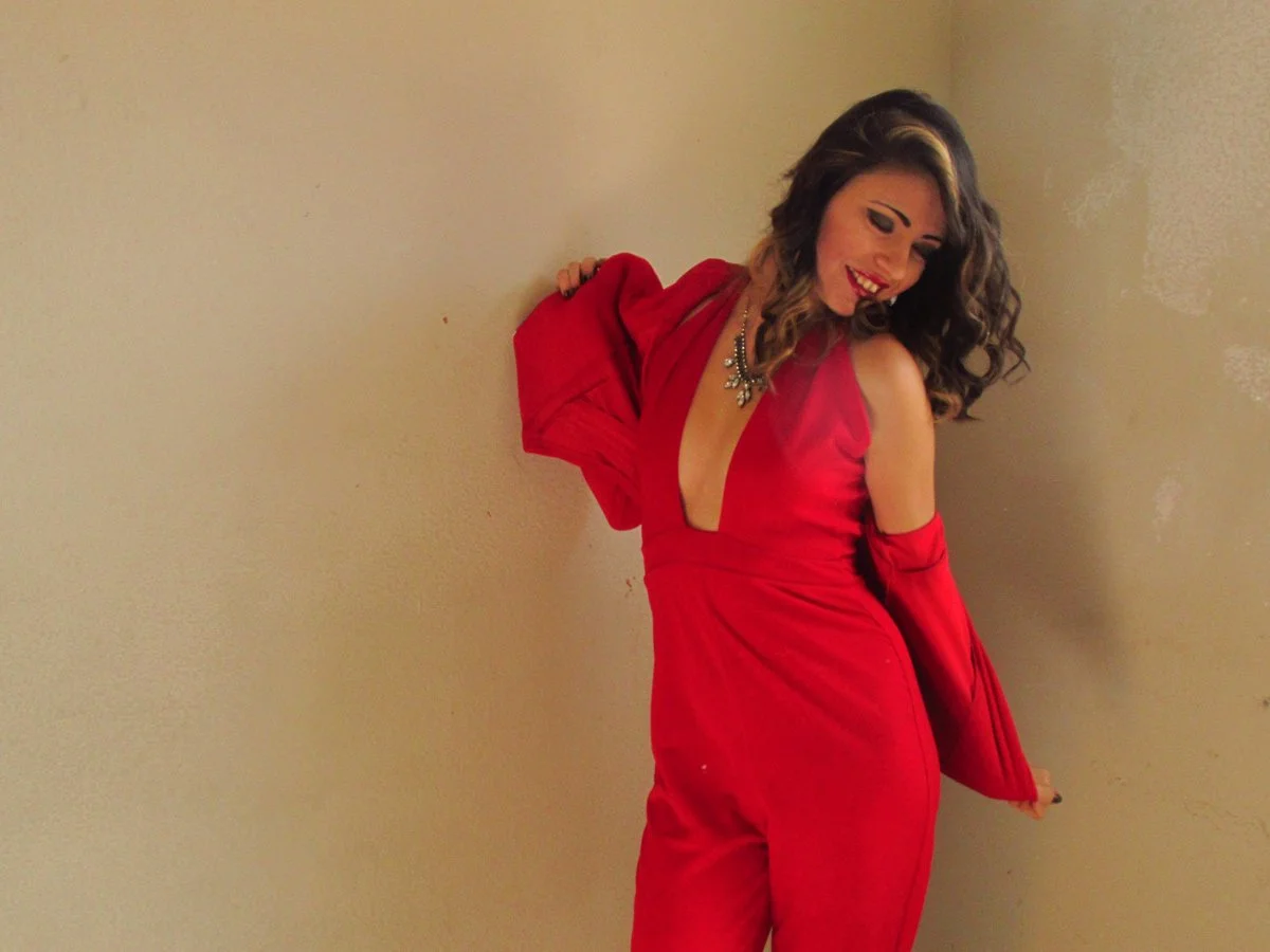 Woman wearing a red dress with a deep neckline, smiling and looking down, with curly hair and a statement necklace, standing against a beige wall.