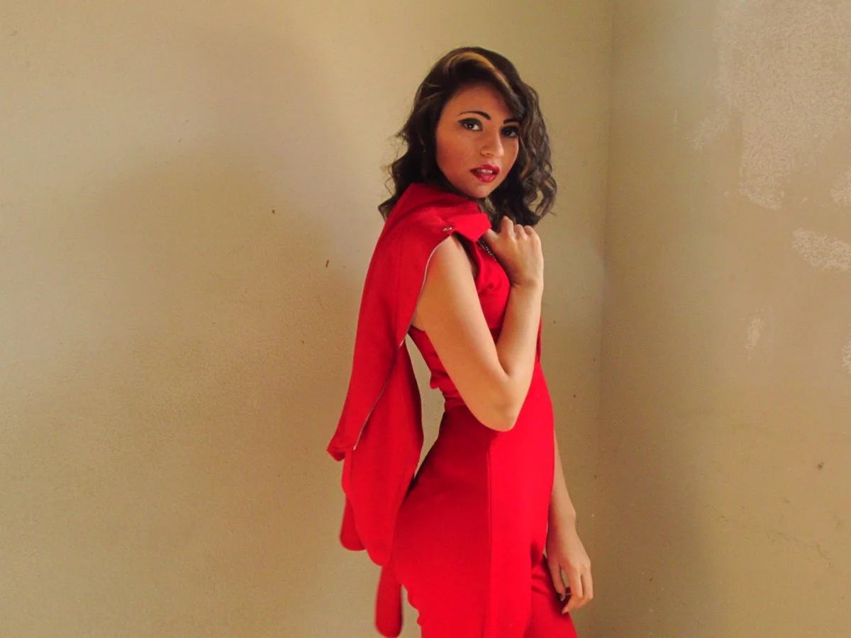 Young woman with dark brown hair and makeup wearing a red dress and a red jacket over her shoulder, standing against a beige wall.