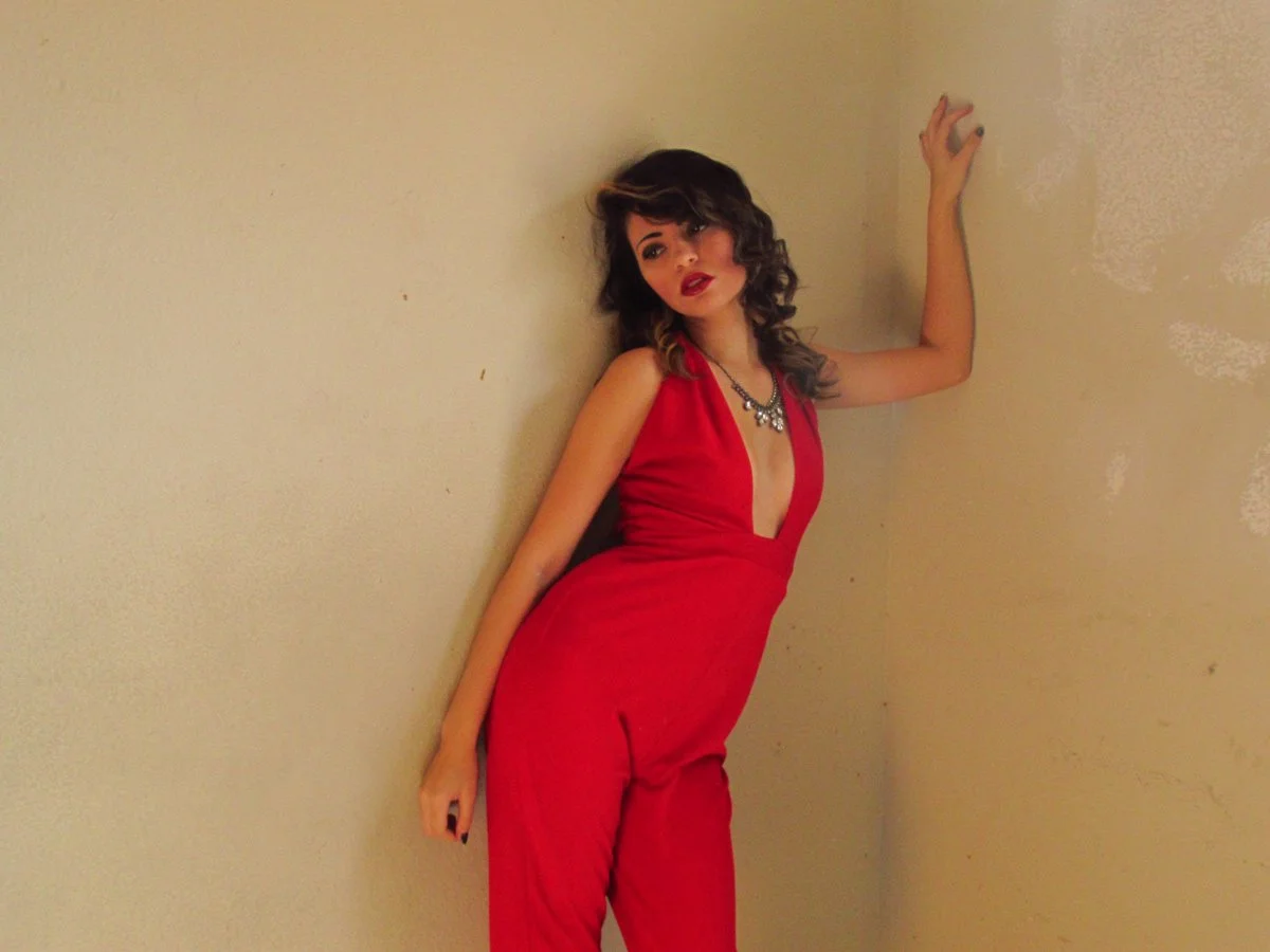 A woman in a red sleeveless jumpsuit with a plunging neckline, posing against a beige wall with her left hand touching the wall and her right hand hanging down, wearing a necklace and red lipstick.