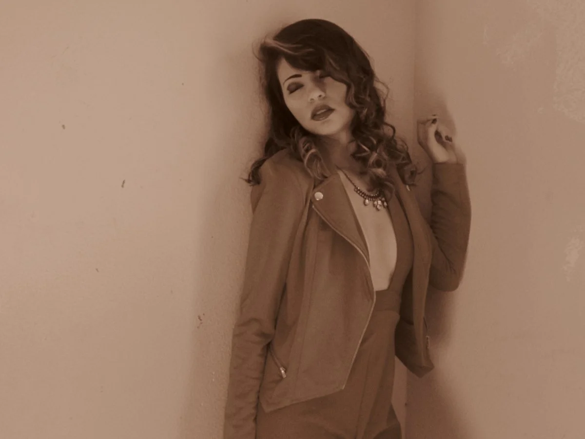 A woman with curly hair and makeup posing against a beige wall, wearing a brown jacket, skirt, and jewelry.