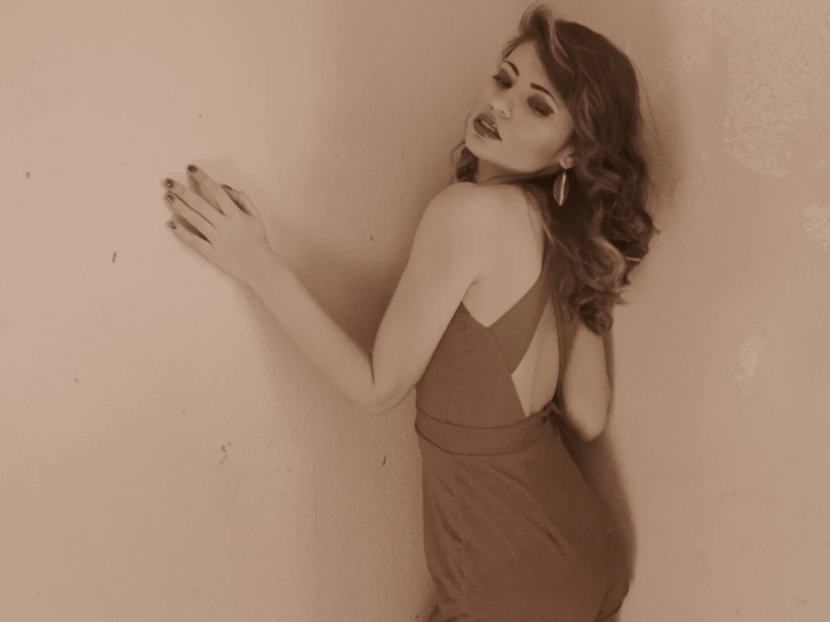 A woman with red hair in curls, wearing a backless dress, leaning against a beige wall with her eyes partially closed and lips slightly parted.