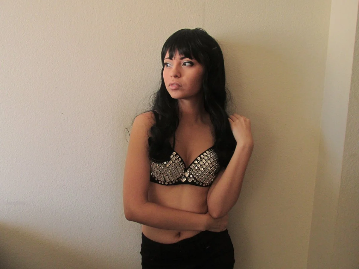 Young woman with long black hair and bangs, wearing a studded black bra and black pants, standing against a beige wall, looking to her left with a serious expression.