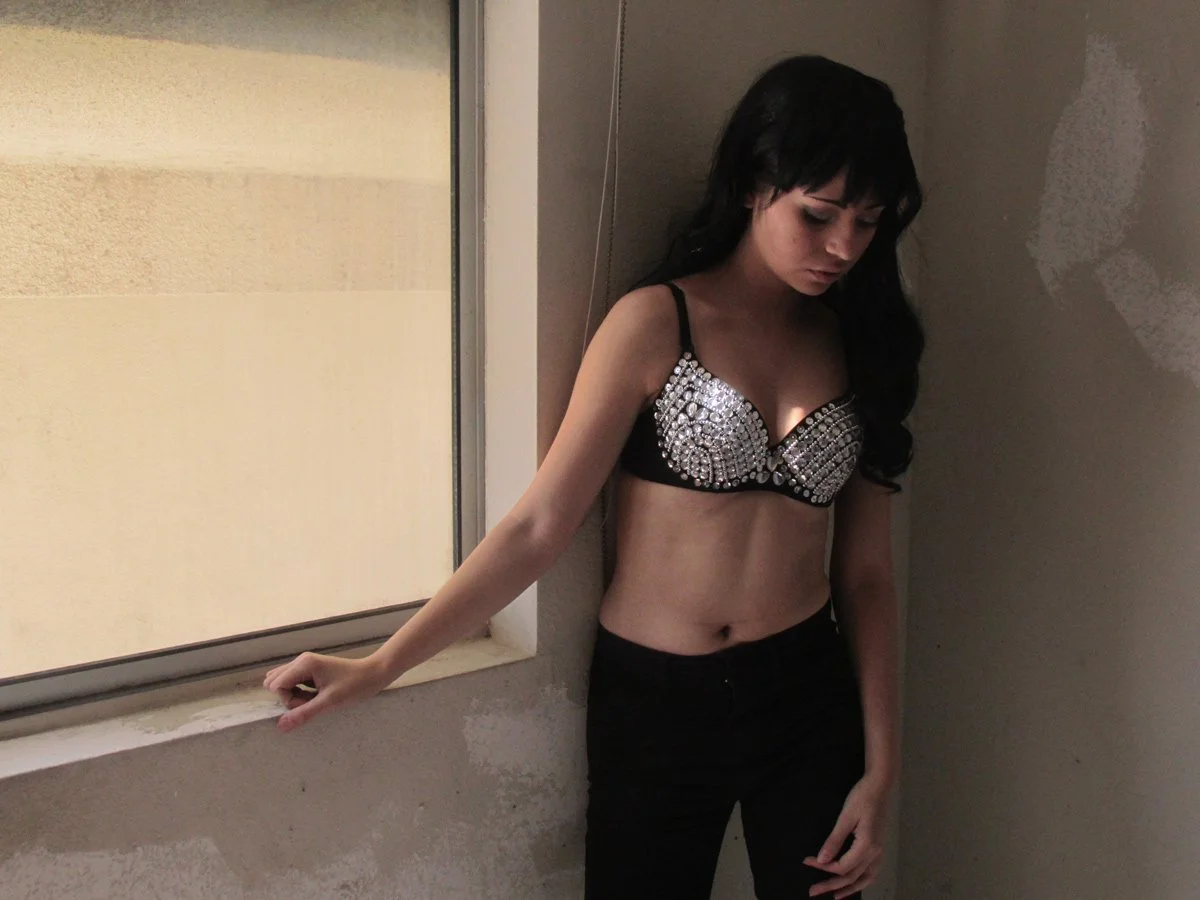 A woman with long black hair is standing in a corner by a window, wearing a black top with shiny embellishments and black pants, looking down with her hand on the window sill.