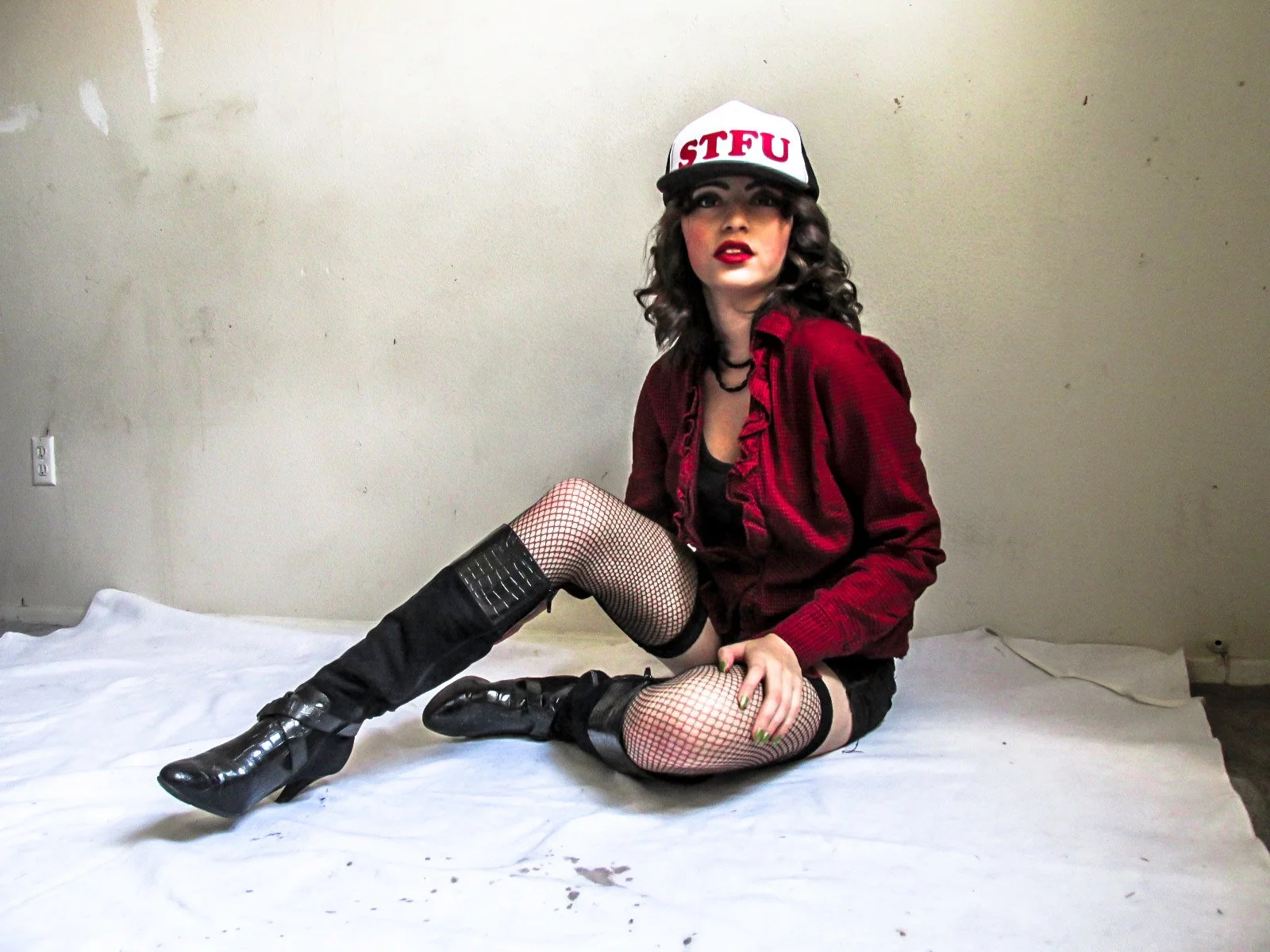 A young woman with curly brown hair and red lipstick, wearing a white cap with red letters reading 'STFU', a red jacket, fishnet stockings, and black knee-high combat boots, sitting on the floor against a plain beige wall.