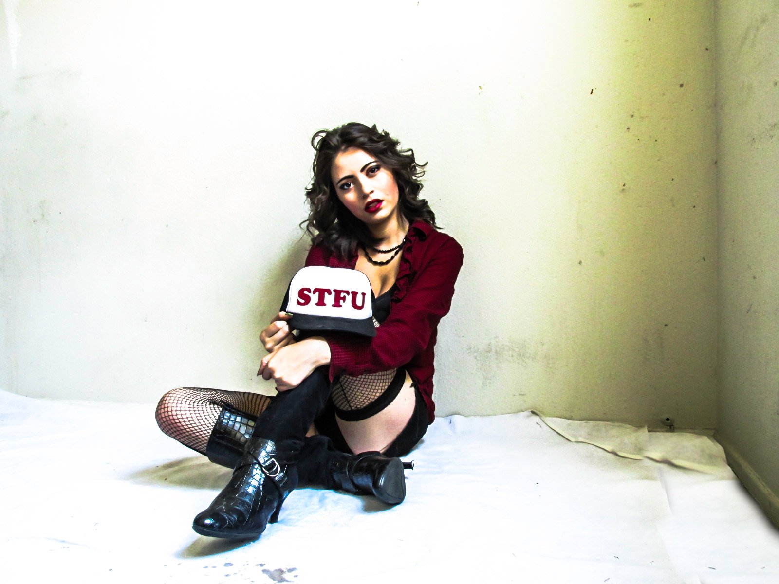 A young woman with dark curly hair and makeup, sitting on the floor against a plain beige wall. She wears fishnet stockings, a red jacket, black boots, and holds a white cap with red letters reading 'STFU.'