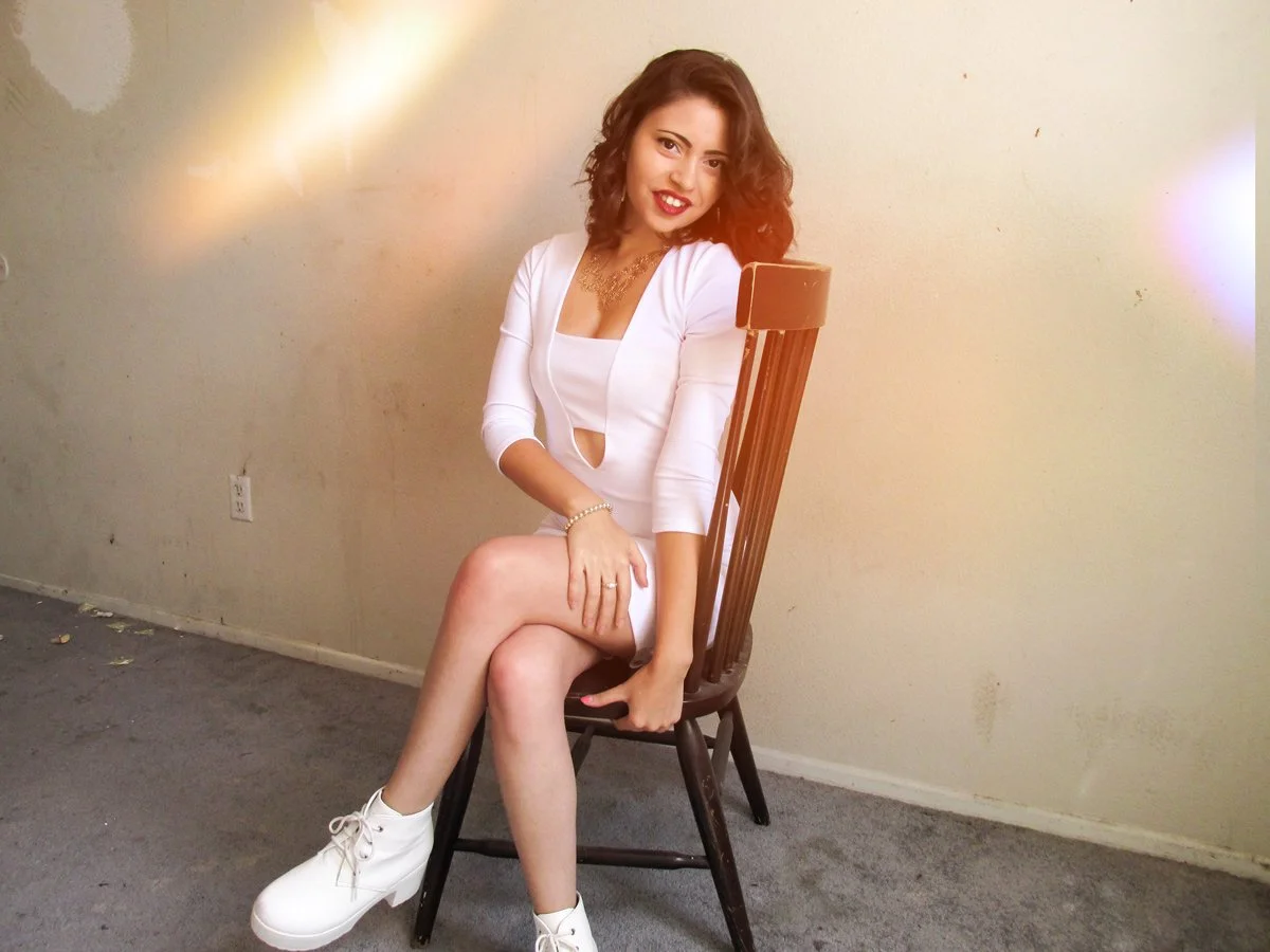 Young woman sitting on a wooden chair in a room with beige walls, wearing a white dress and white shoes, smiling and posing for the camera.