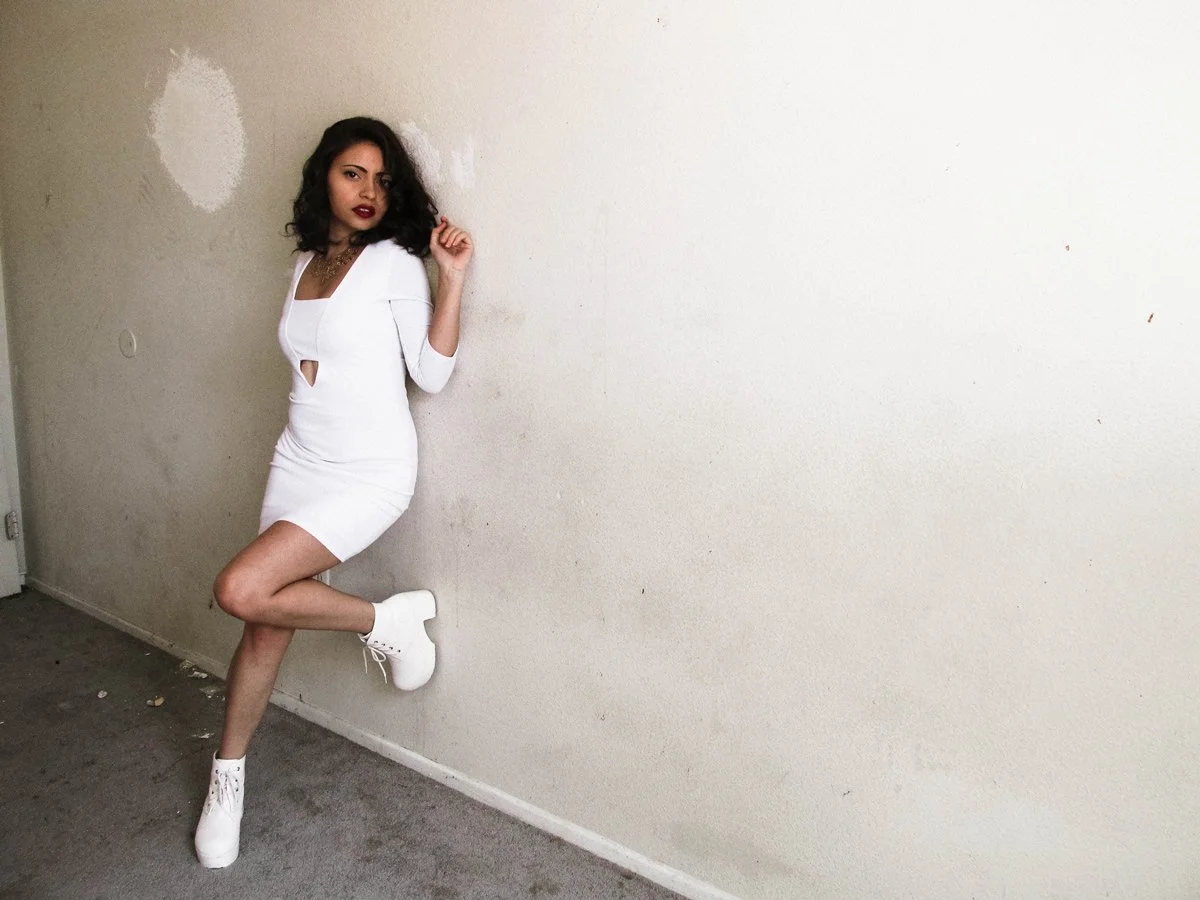 A woman in a white dress and white high-heeled boots standing against a plain, off-white wall, with her left leg bent and foot resting against the wall.
