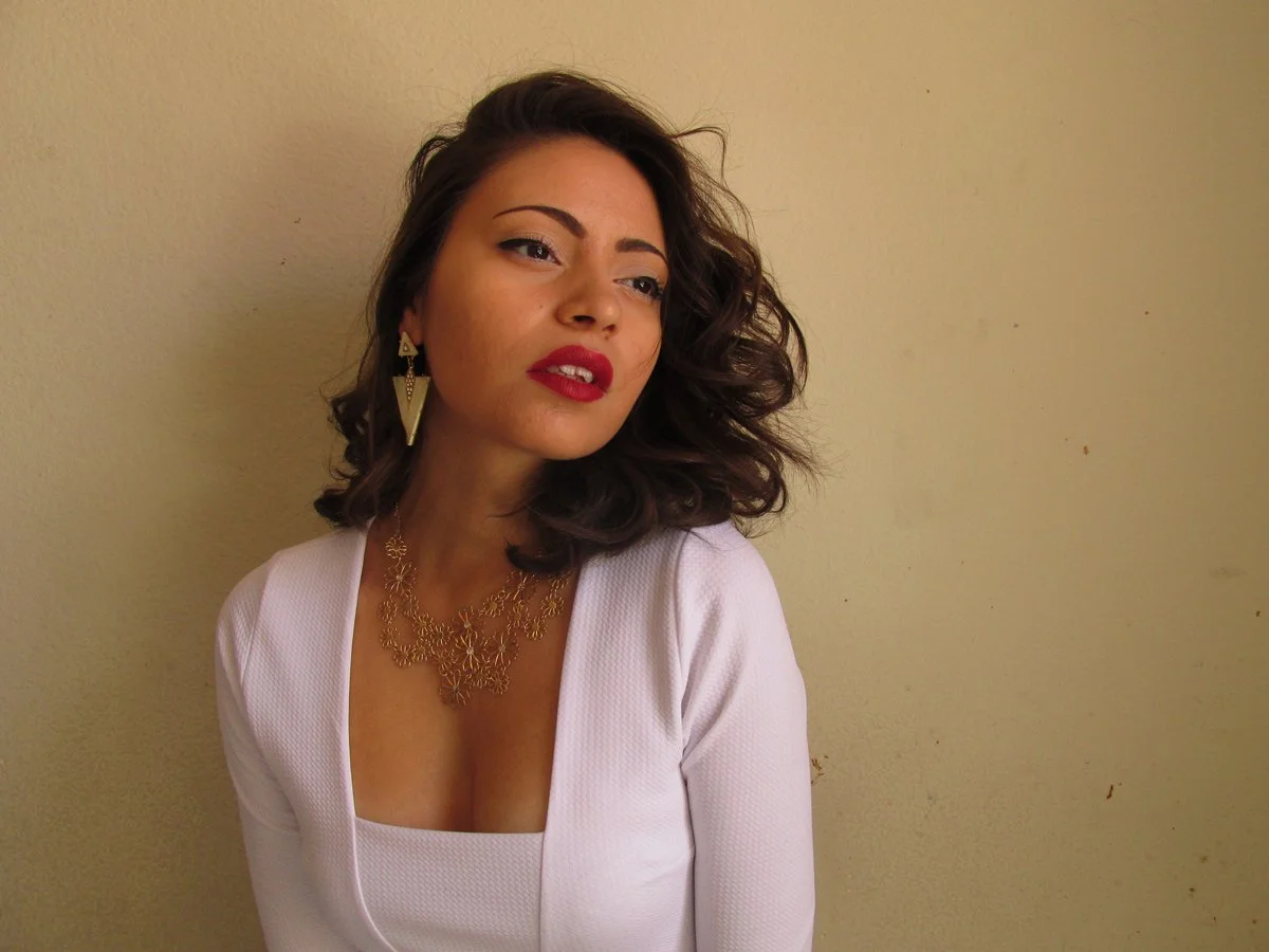A woman with dark, curly hair wearing a white top and gold jewelry, standing against a beige wall.