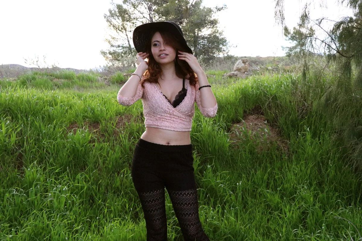 A young woman standing outdoors in a grassy area with trees in the background, wearing a large black hat, a pink lace crop top with black lace underneath, and black lace leggings.