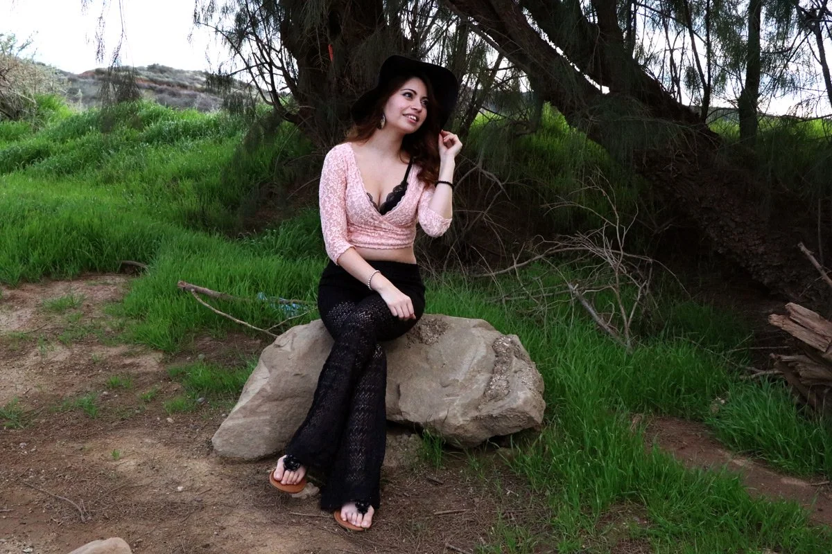 A woman wearing a pink top, black pants, and a black hat sitting on a large rock in a green outdoor setting with trees and grass.