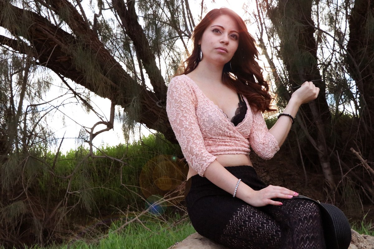Young woman with long wavy hair sitting outdoors near trees, wearing a pink lace top, black patterned tights, and jewelry, looking at the camera with a serious expression.