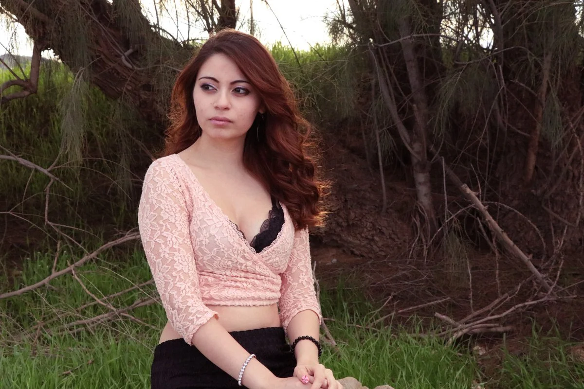 A young woman with long, wavy auburn hair sitting outdoors on grass, wearing a peach lace crop top with black inner lining, a black skirt, and jewelry, with trees and a natural background.