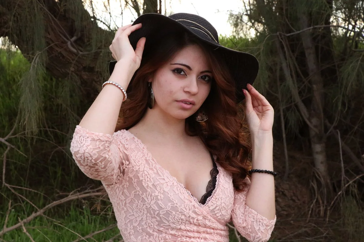 A young woman with long, wavy brown hair wearing a pink lace dress and a black wide-brimmed hat, holding the brim of the hat with both hands, standing outdoors with trees and greenery in the background.