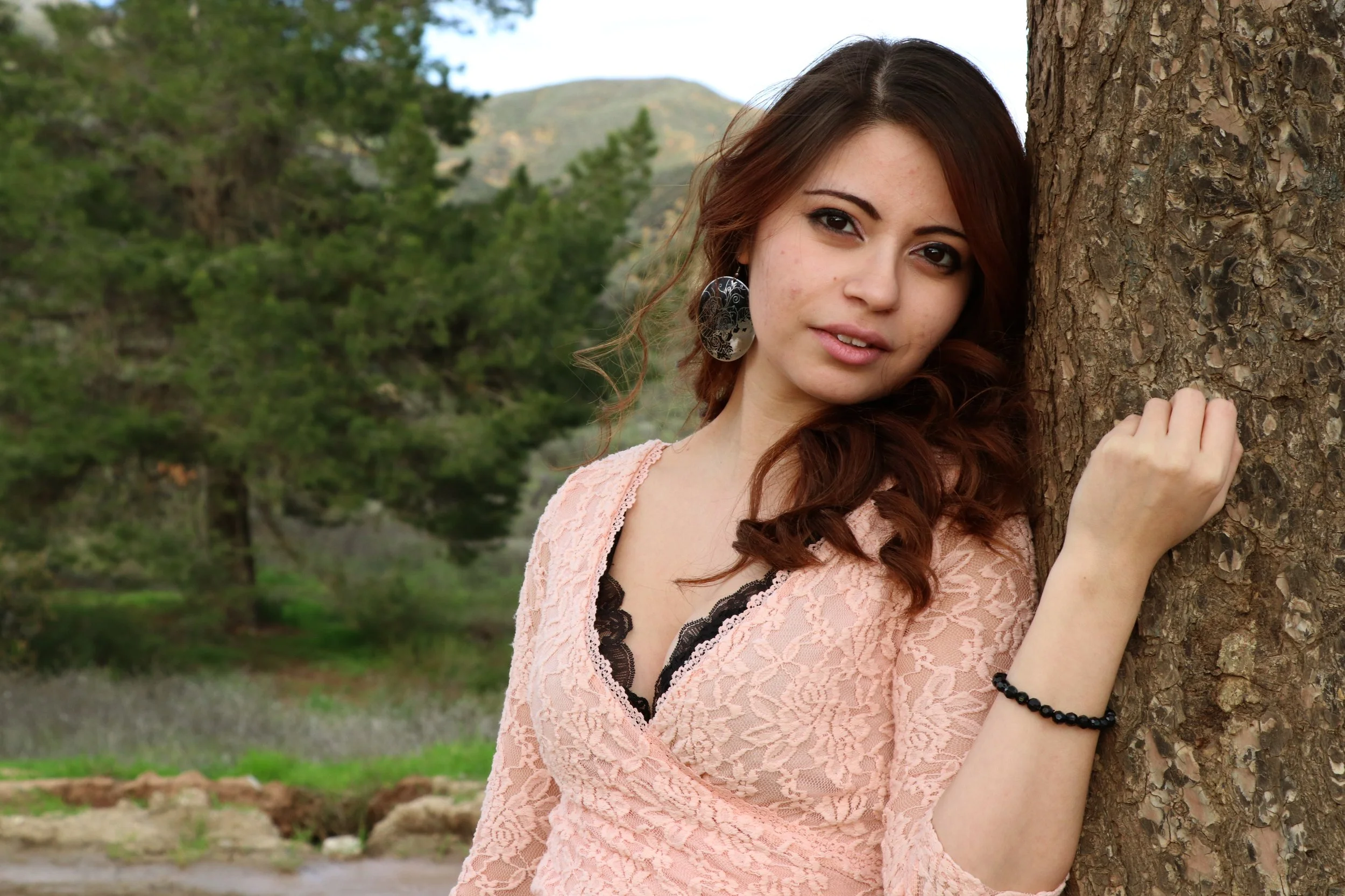 A young woman with brown hair and large earrings leaning against a tree in a scenic outdoor setting with trees and mountains in the background.