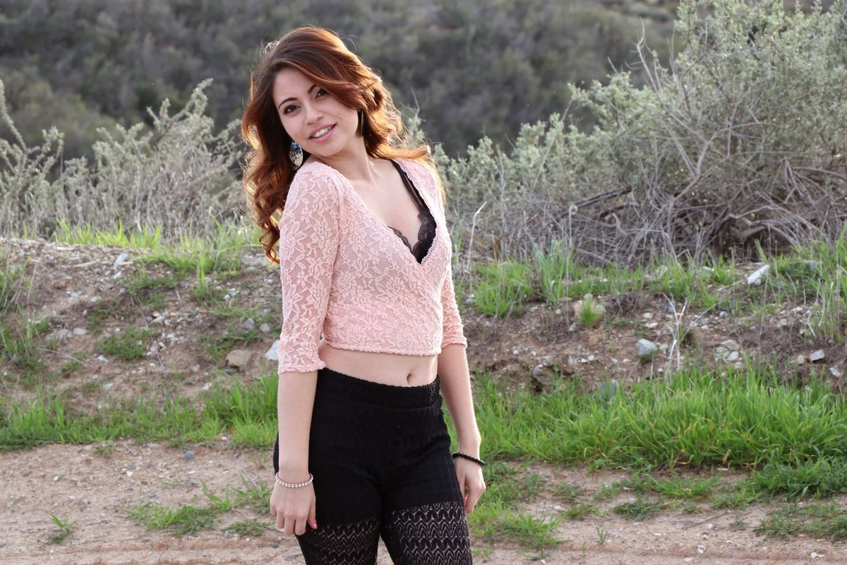 A young woman with curly, reddish-brown hair standing outdoors in a grassy and rocky area with bushes and trees in the background, smiling at the camera.