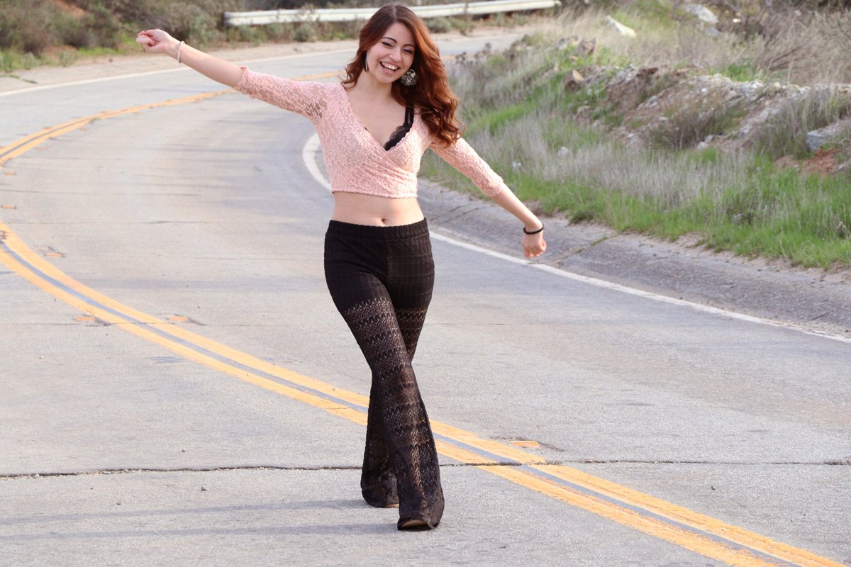 A young woman with red hair walking on a curved rural road, smiling with arms outstretched.