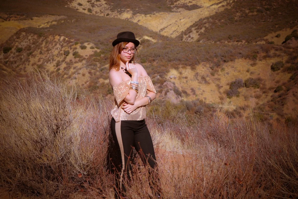 A young woman with long brown hair, glasses, and a hat, standing in a dry, hilly landscape with brown and purple vegetation.
