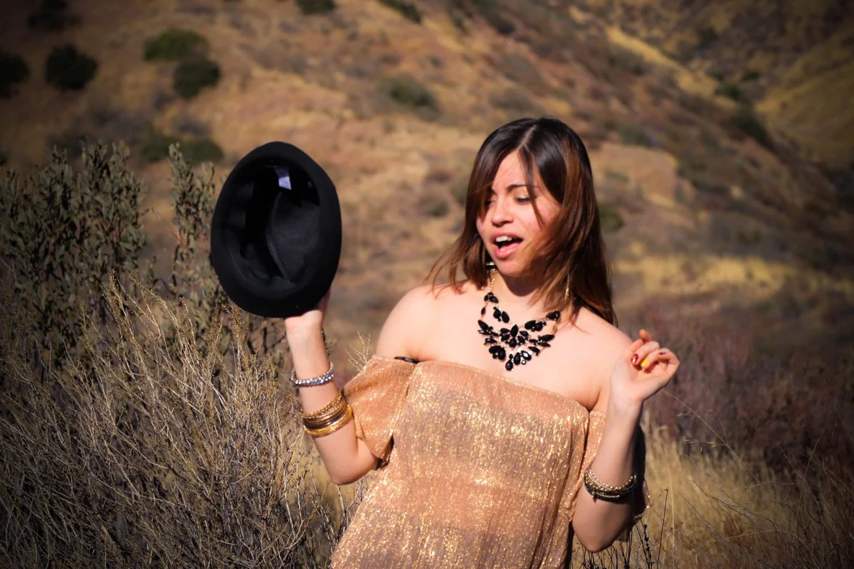 A young woman with shoulder-length brown hair wearing a shiny gold off-shoulder dress, holding a black fedora hat in one hand and dancing outdoors with a scenic mountainous background.
