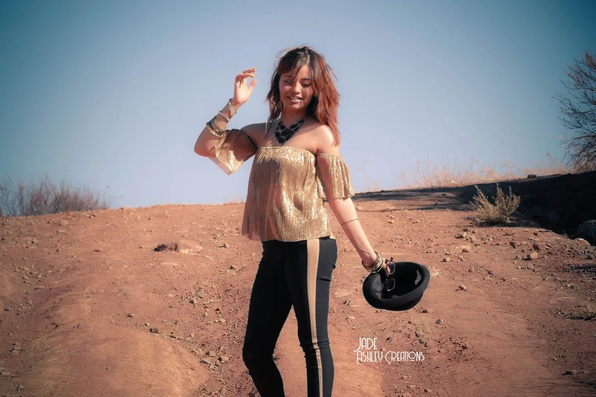 A woman standing on a dirt hill holding a black hat, wearing a metallic gold off-the-shoulder top, black pants with a gold stripe, and jewelry, with a clear blue sky in the background.