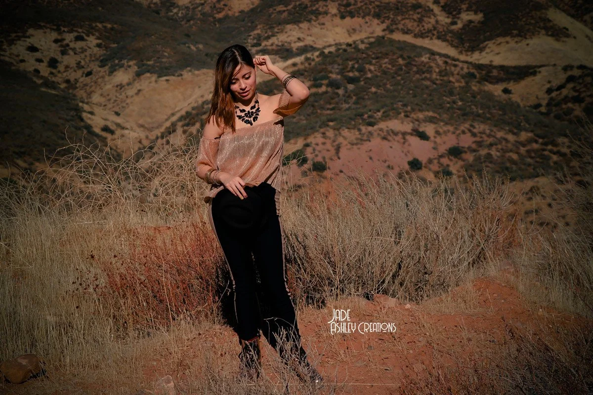 A young woman standing in tall, dry grass in a desert landscape with rolling hills in the background, wearing a shiny gold off-shoulder top, black pants, and jewelry.