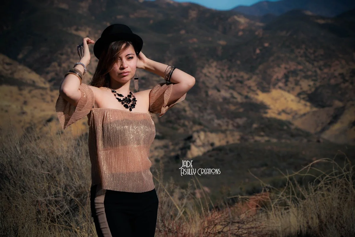 A woman in a gold-colored top and black pants posing outdoors in a mountainous landscape, wearing a black hat and jewelry.