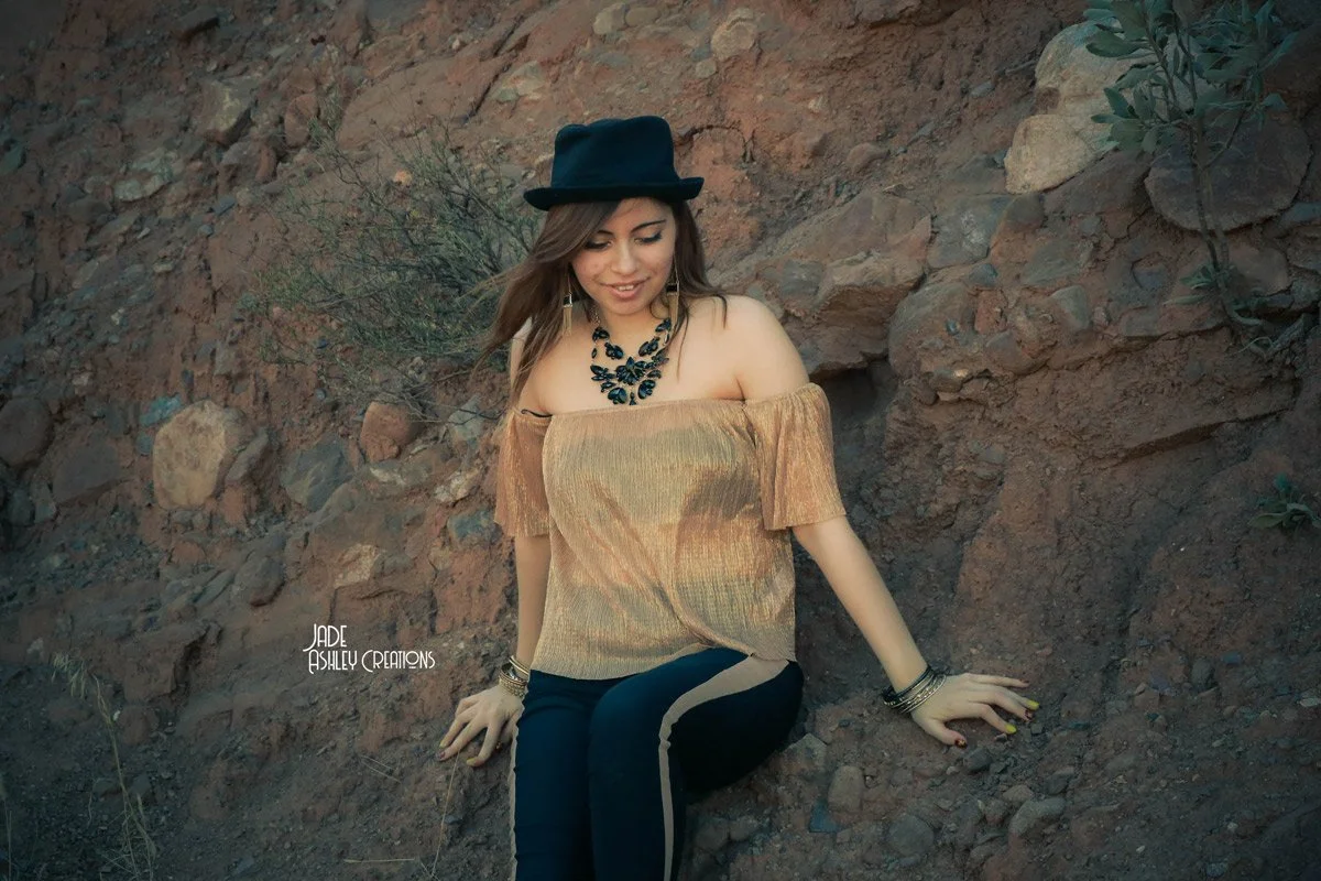 A woman wearing a black hat, gold off-the-shoulder blouse, and black pants with a beige side stripe, sitting outdoors against a rocky background, smiling and looking down.