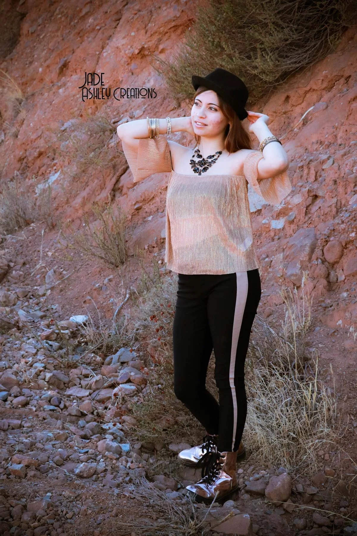 A young woman standing outdoors on rocky terrain with dry bushes, wearing a metallic off-the-shoulder top, black pants with a beige stripe, and metallic boots, accessorized with bracelets, necklace, and a black hat.