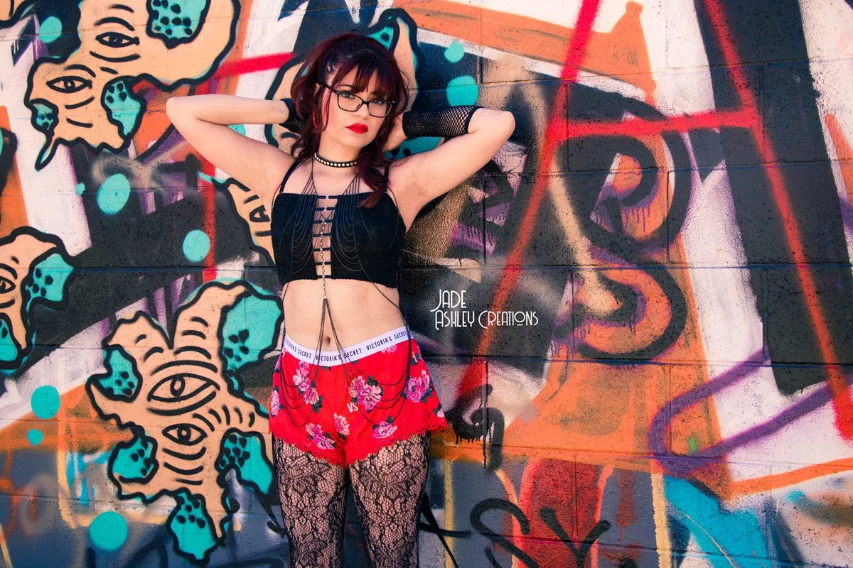 Young woman with glasses and dark hair posing in front of colorful graffiti wall, wearing a black lace top, red floral shorts with chains, lace leggings, and fishnet gloves.