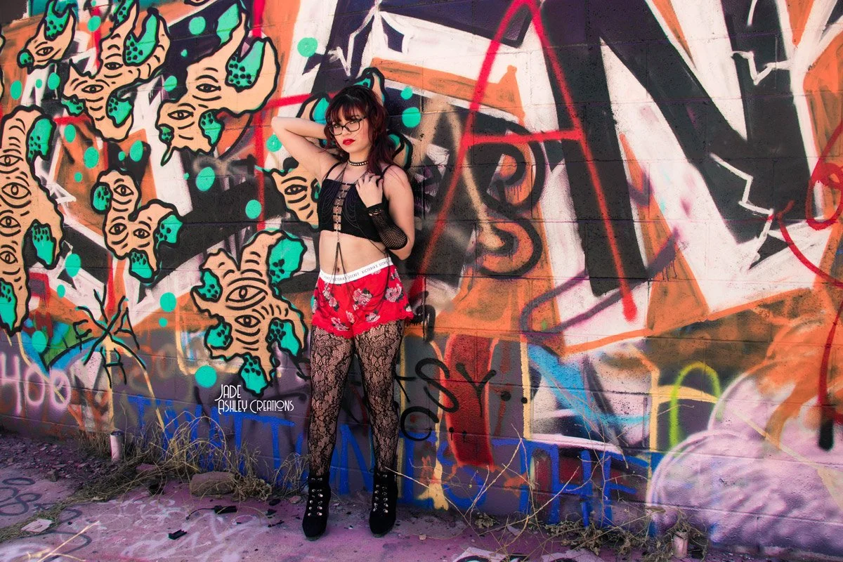 Young woman with dark hair, glasses, and red lipstick standing in front of a colorful graffiti wall, wearing a black mesh top, red floral shorts, patterned fishnet stockings, and platform shoes.