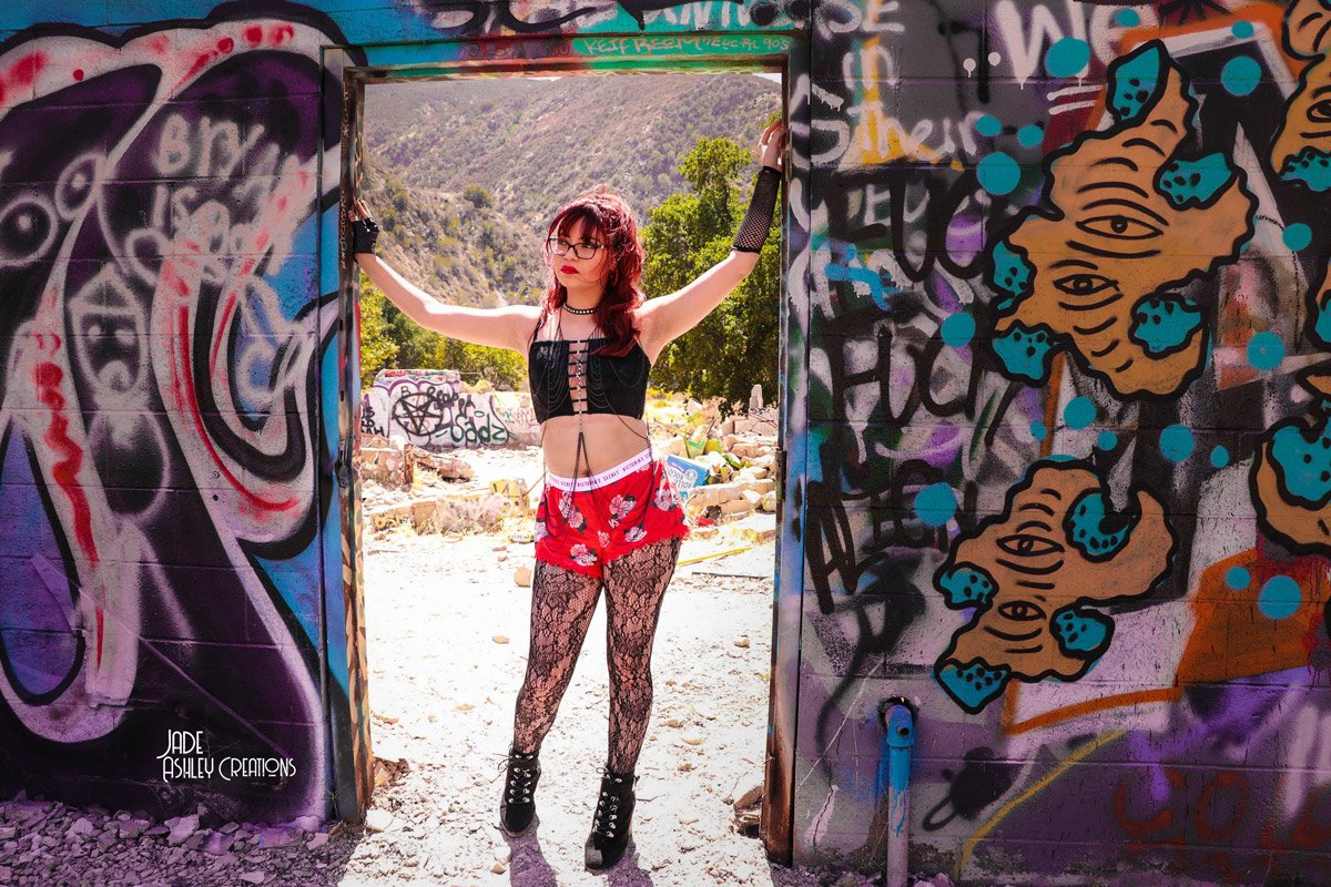A young woman with red hair, glasses, and a punk style outfit, standing in a graffiti-covered doorway of an abandoned building in a desert landscape.