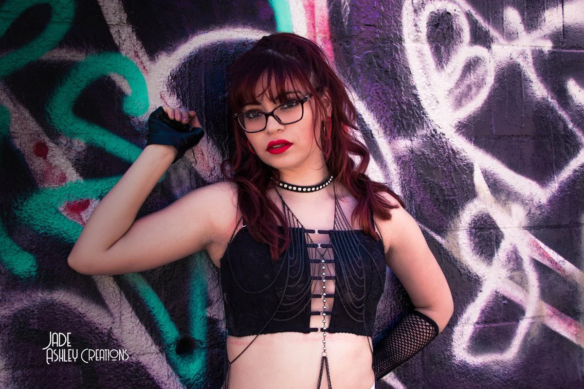 A young woman with red hair, glasses, and red lipstick standing against a graffiti-covered wall, wearing a black lace top, fishnet gloves, and jewelry.