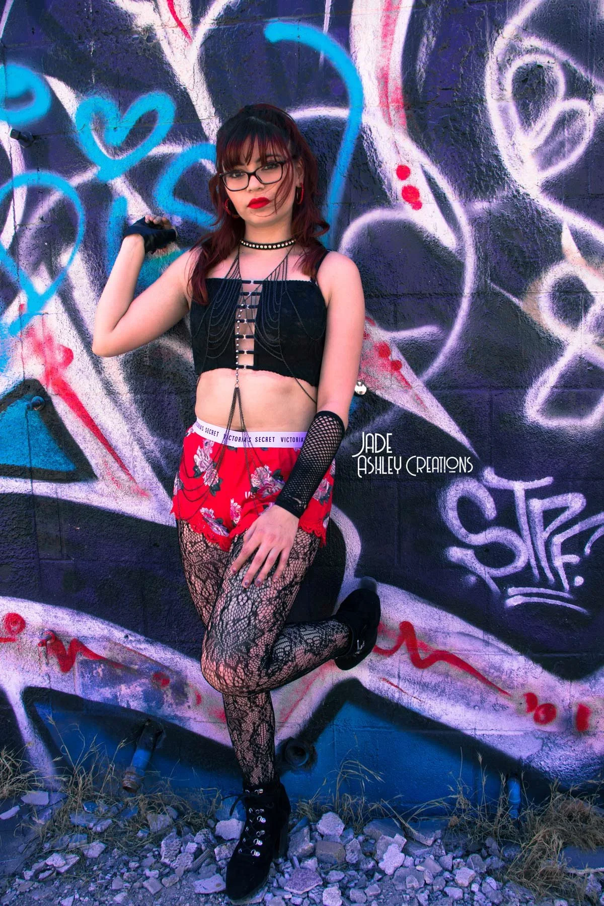 A young woman with red hair, glasses, and red lipstick, posing against a graffiti-covered wall, wearing a black crop top, red floral shorts, fishnet tights, and black shoes, accessorized with jewelry and fishnet arm sleeves.