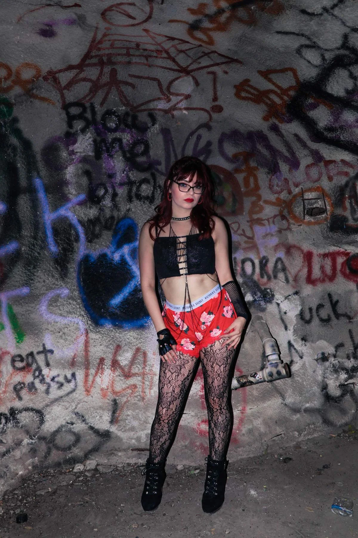 A woman with dark hair, glasses, and red lipstick standing in front of a graffiti-covered wall. She is wearing a black lace top, red floral shorts, black lace tights, black platform boots, and accessories including gloves, necklaces, and earrings.