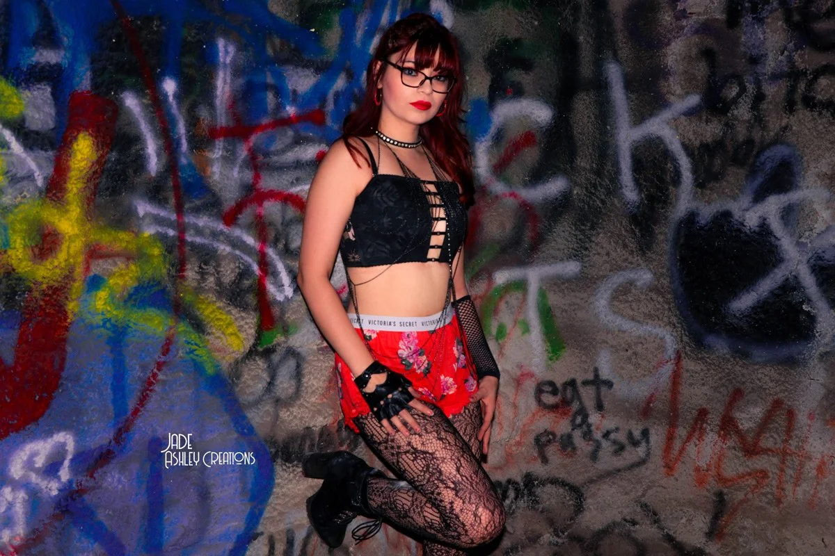 Young woman with red hair, glasses, and tattoos wearing a black lace crop top, red floral shorts, fishnet stockings, and black boots, standing in front of graffiti-covered wall.