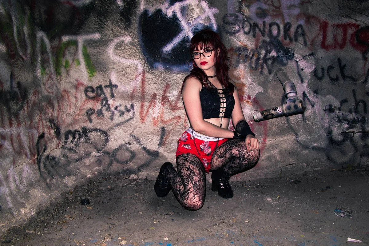 A young woman with dark hair and glasses squats in front of a graffiti-covered wall, wearing a black top, red shorts with floral pattern, lace tights, and black boots.