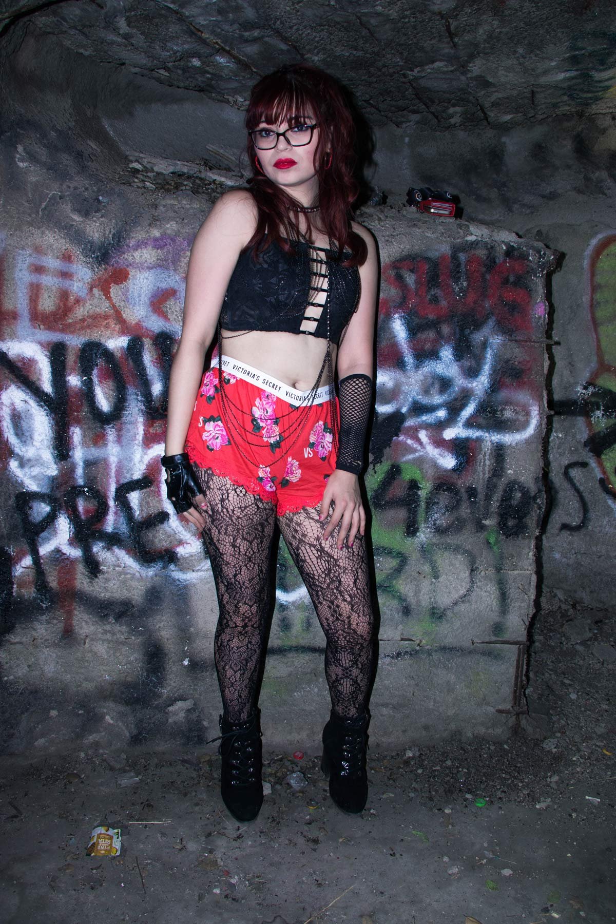A woman with red hair, glasses, and gothic style clothing standing against a graffiti-covered wall in a dark, underground setting.