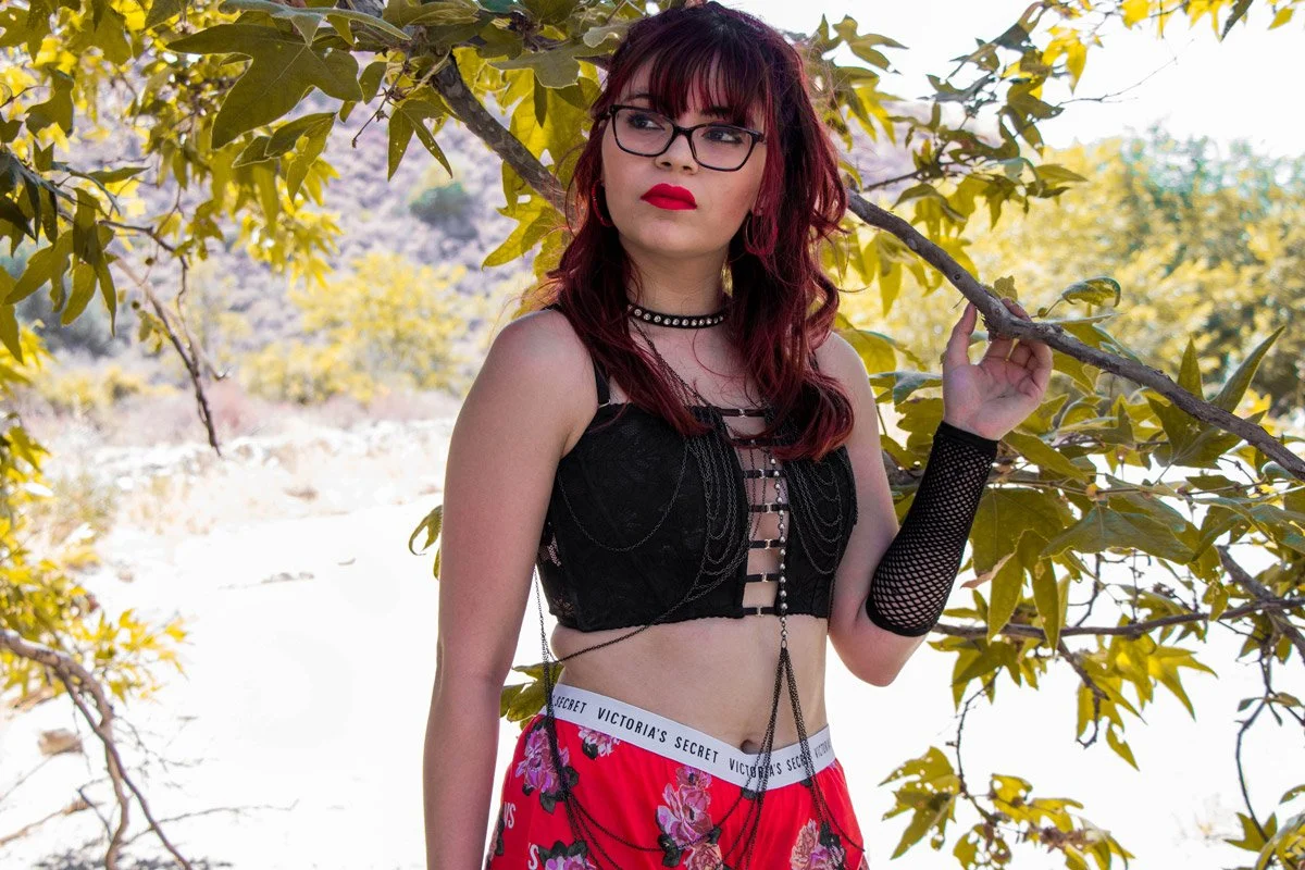 A young woman with red hair and glasses standing outdoors under a tree, wearing a black lace top, red patterned shorts, and arm warmers, holding a tree branch with one hand.