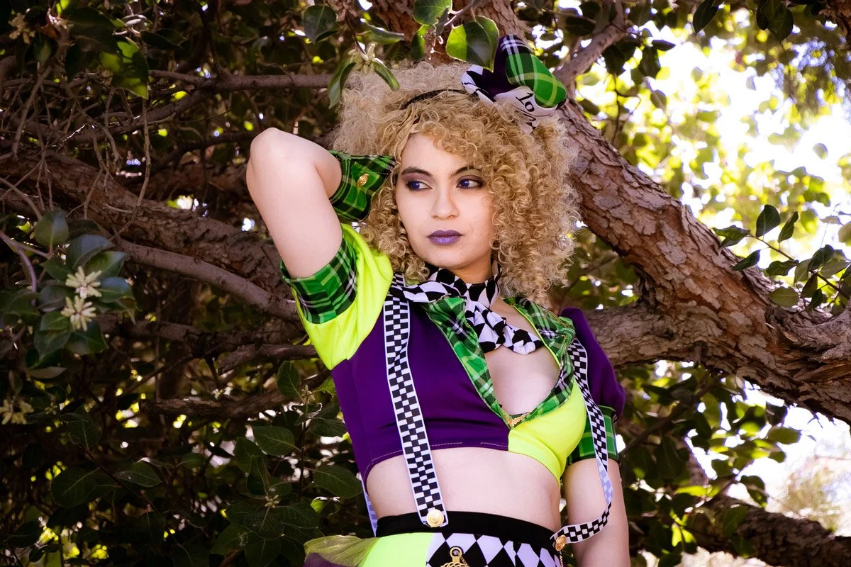 Young woman with curly blonde hair and purple lipstick wearing a colorful outfit with green, purple, and black patterns, standing in front of a tree with branches and leaves, looking to the side with one arm raised.