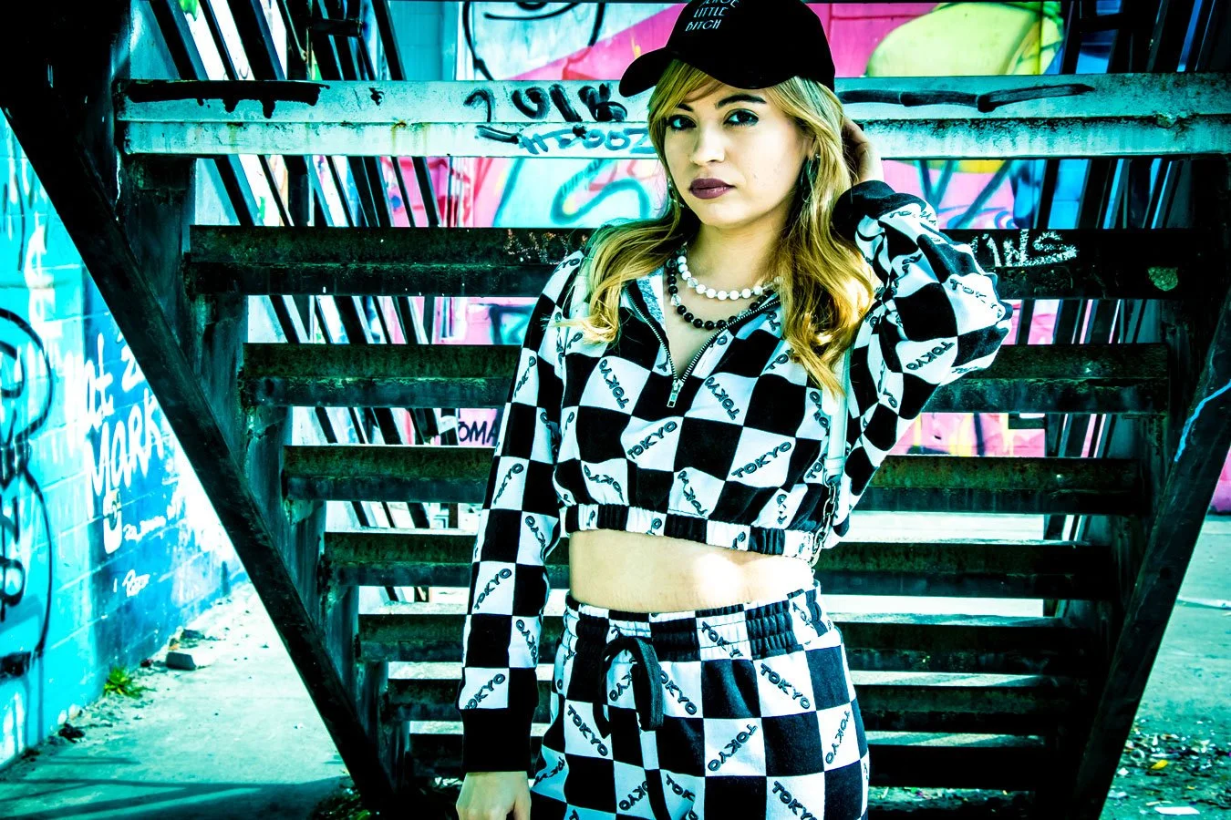 Young woman with blonde hair in checkered Tokyo-themed outfit standing in front of graffiti-covered urban fire escape.