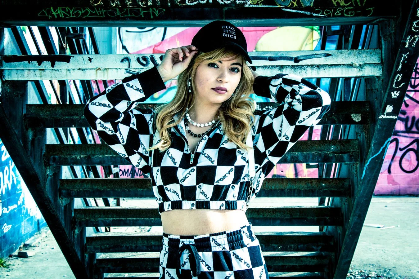 Young woman with blonde hair wearing a black cap, checkered jacket, and matching pants, standing in front of a wooden staircase with graffiti in the background.