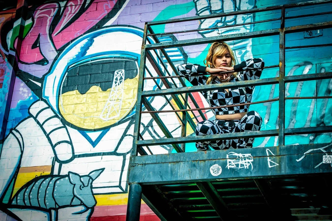 A woman sits on a metal fire escape with her arms resting on the railing, in front of a colorful graffiti mural of an astronaut helmet with a gold reflection visor and a rainbow backdrop.