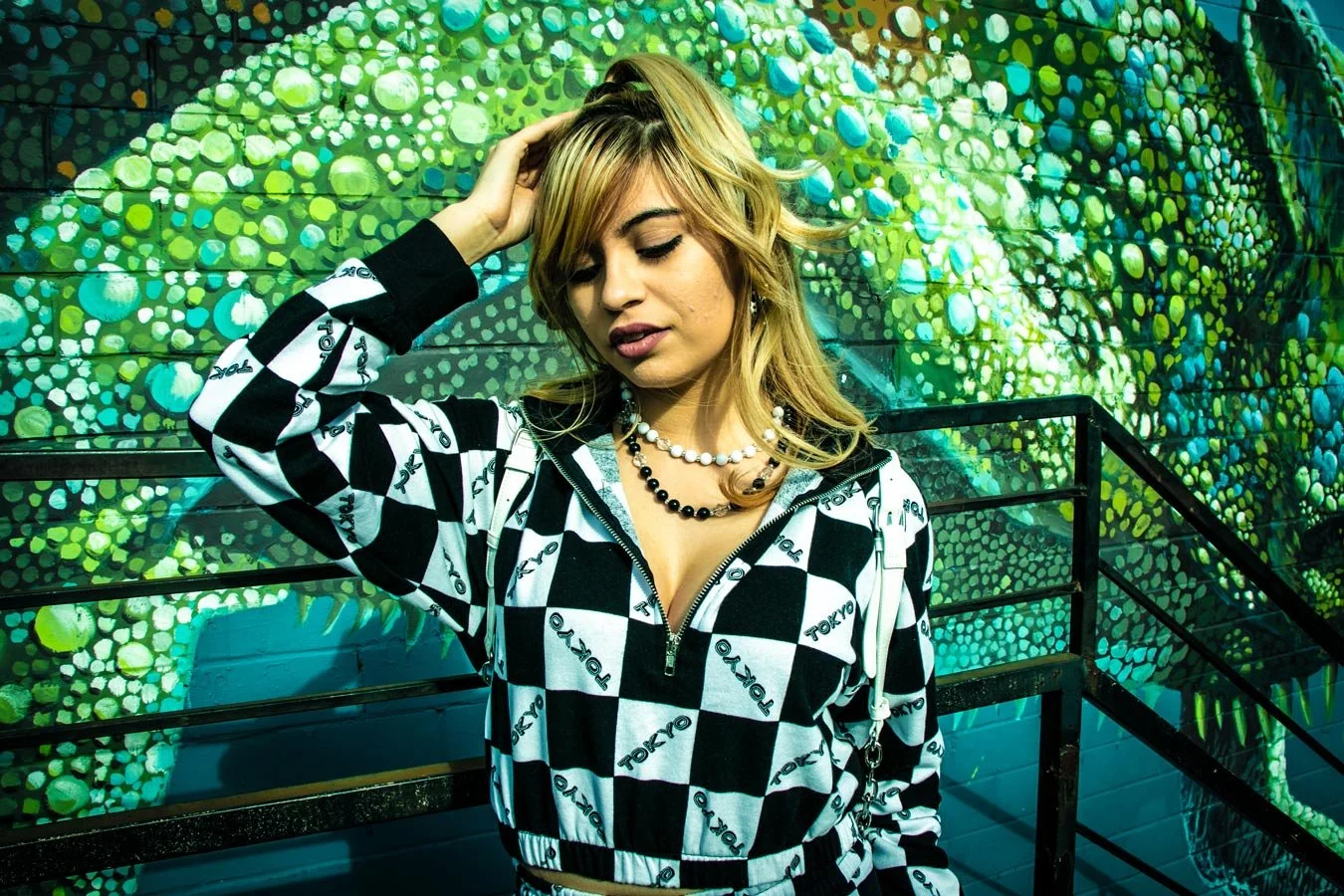A young woman with blonde hair and layered necklaces, wearing a black and white checkered jacket with 'TOKYO' text, standing in front of a colorful mural with green, yellow, and blue circular patterns.