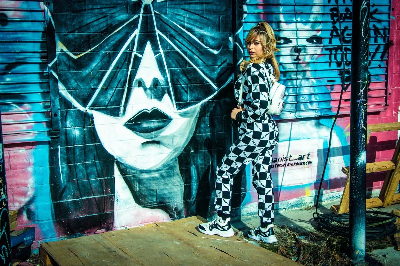 A woman wearing a black and white checkered outfit and sneakers stands next to a graffiti mural of a woman's face with glasses on a brick wall.