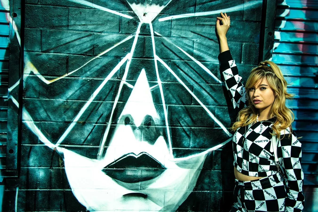 A woman with blonde hair and a checkered black and white jacket and pants, standing next to a large street art mural of a woman's face on a brick wall, in an urban environment.
