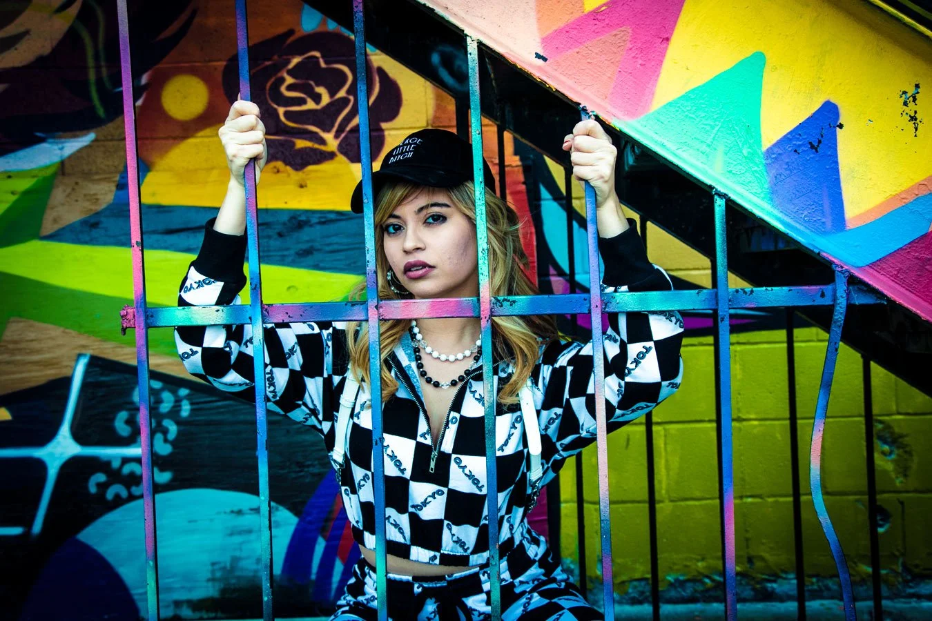 A young woman with blonde hair, wearing a black cap, black and white checker patterned outfit, and layered necklaces, holding onto metal bars in front of a colorful graffiti mural background.