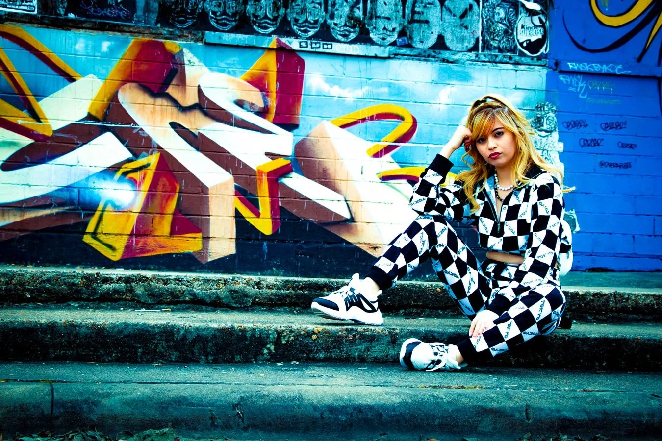 A young woman sitting on concrete steps next to a colorful graffiti mural on a brick wall. She is wearing a black and white checkered outfit and sneakers, and has blond hair with loose waves.