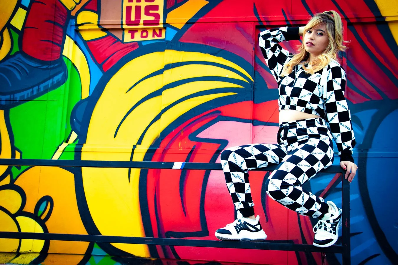 A woman wearing a black and white checkered crop top and matching pants, sitting on a metal railing in front of a colorful graffiti mural that features various bright colors including red, yellow, green, and blue.