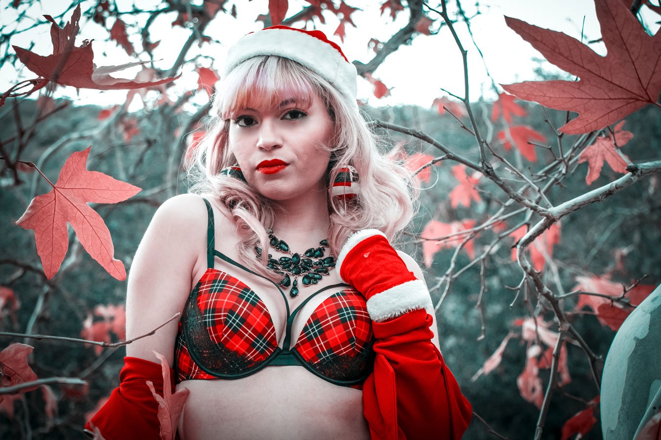 A woman wearing a Santa hat, red and black plaid lingerie top, and red gloves, standing among red autumn leaves on a tree in an outdoor setting.