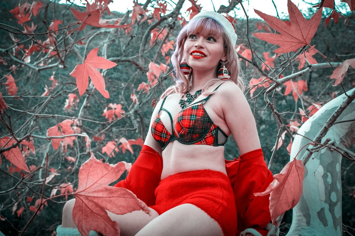 A woman with blonde hair, wearing a Christmas-themed lingerie top, red fuzzy skirt, and red gloves, posing outdoors among red autumn leaves, with a festive hat and jewelry, smiling and looking to the side.
